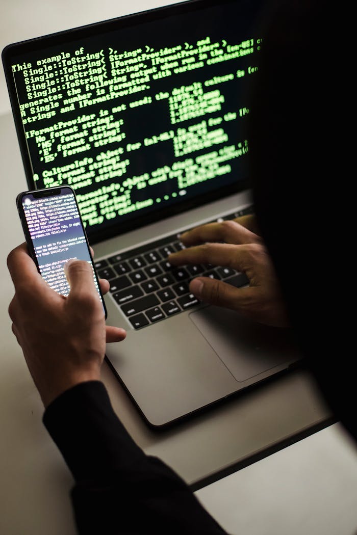 digital A hooded figure engaged in hacking using a laptop and smartphone in low light.